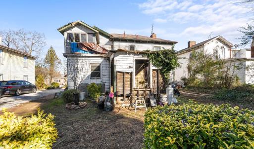 The back of the house. A view of the shared driveway, the sunporch, the shell of a room, kitchen window, a/c unit, plantings.