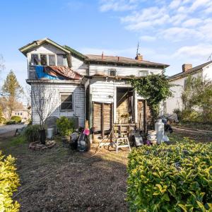 The back of the house. A view of the shared driveway, the sunporch, the shell of a room, kitchen window, a/c unit, plantings.